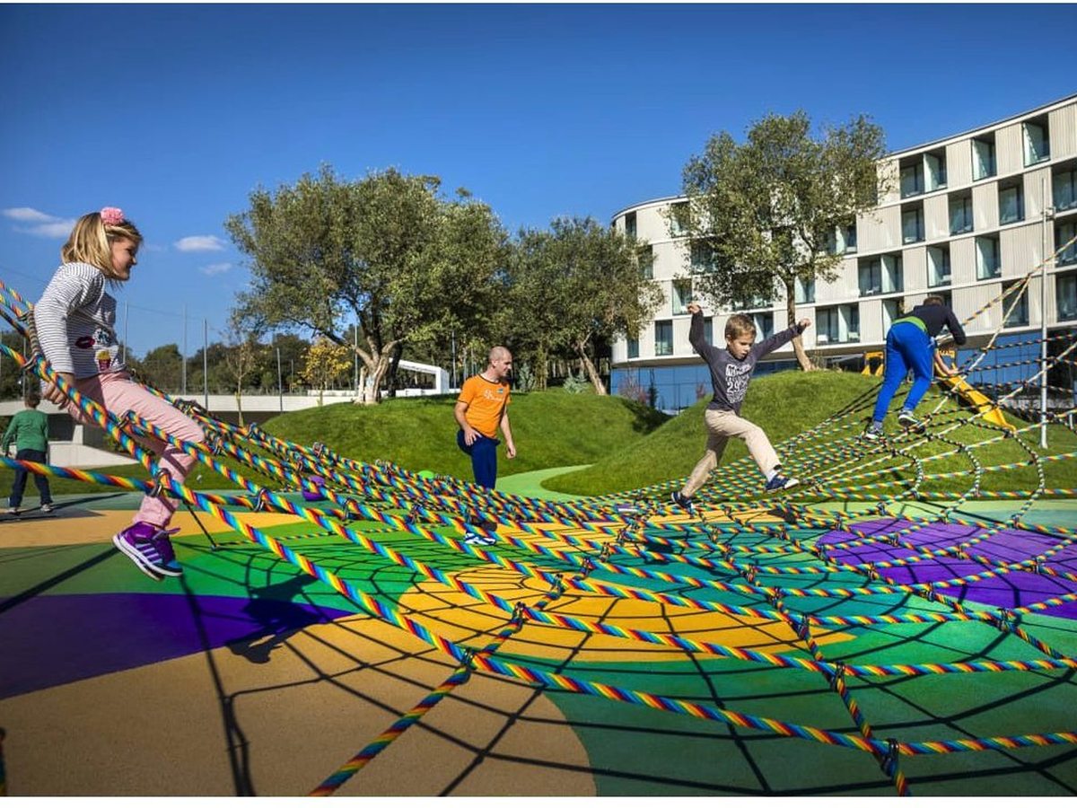 Children crossing a large rope climbing net woven in rainbow-striped ropes, set over green and purple rubber play surfacing outside the hotel.