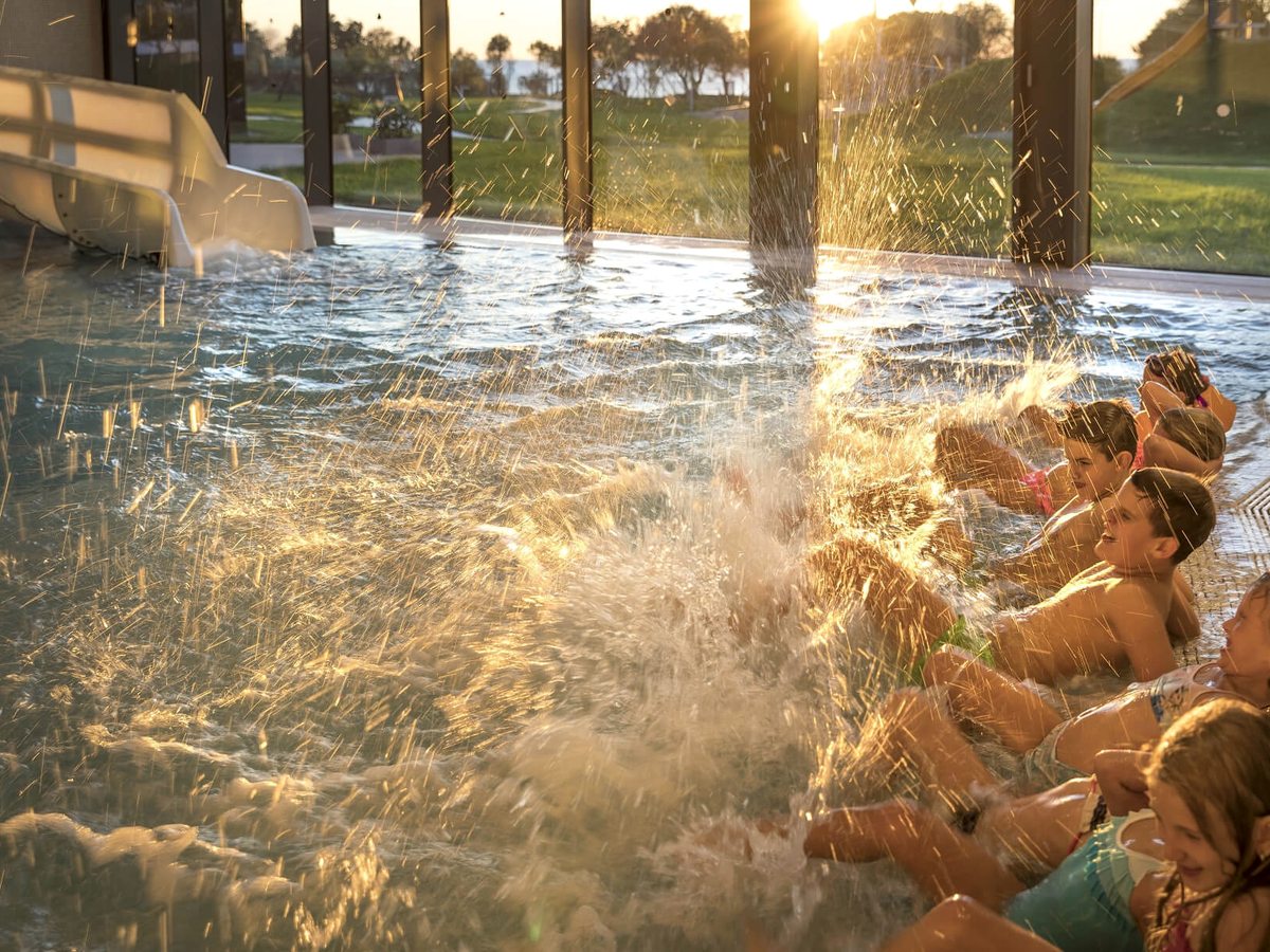 Children sitting along the edge of an indoor pool splashing in the water, a white water slide entering the pool and low sun through full-height windows.