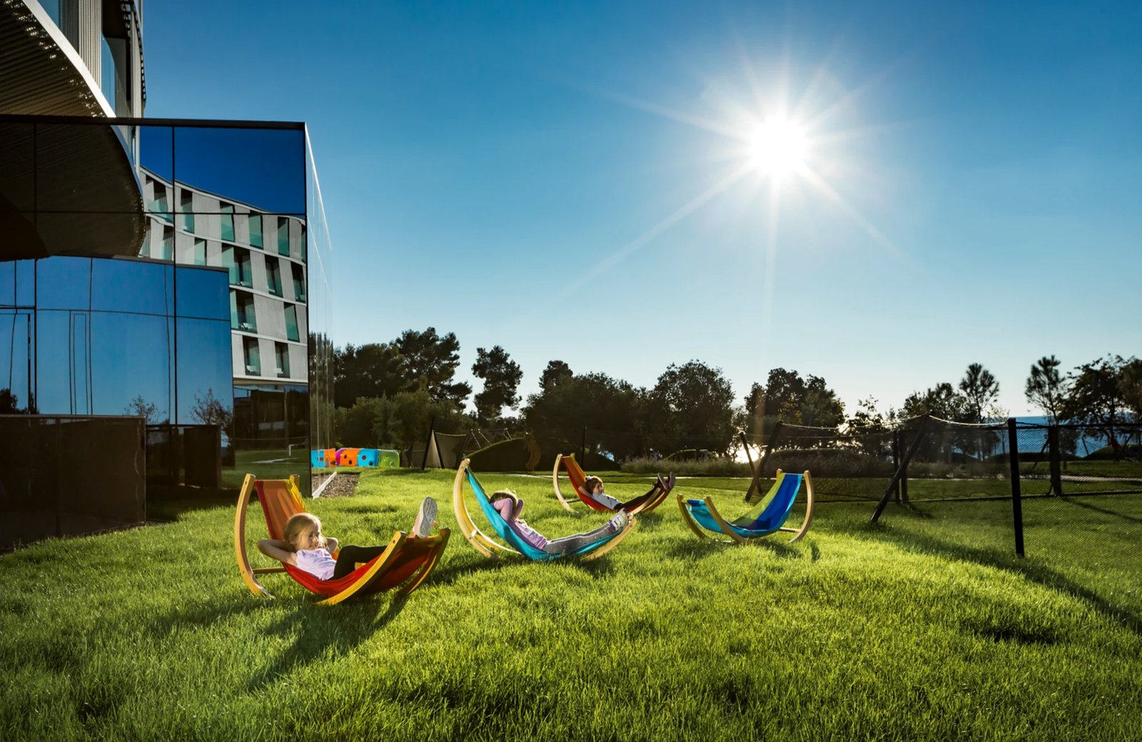 Children lying in brightly coloured hammocks on a sunlit lawn beside the glass-fronted Family Hotel Amarin, with pines and the sea behind.
