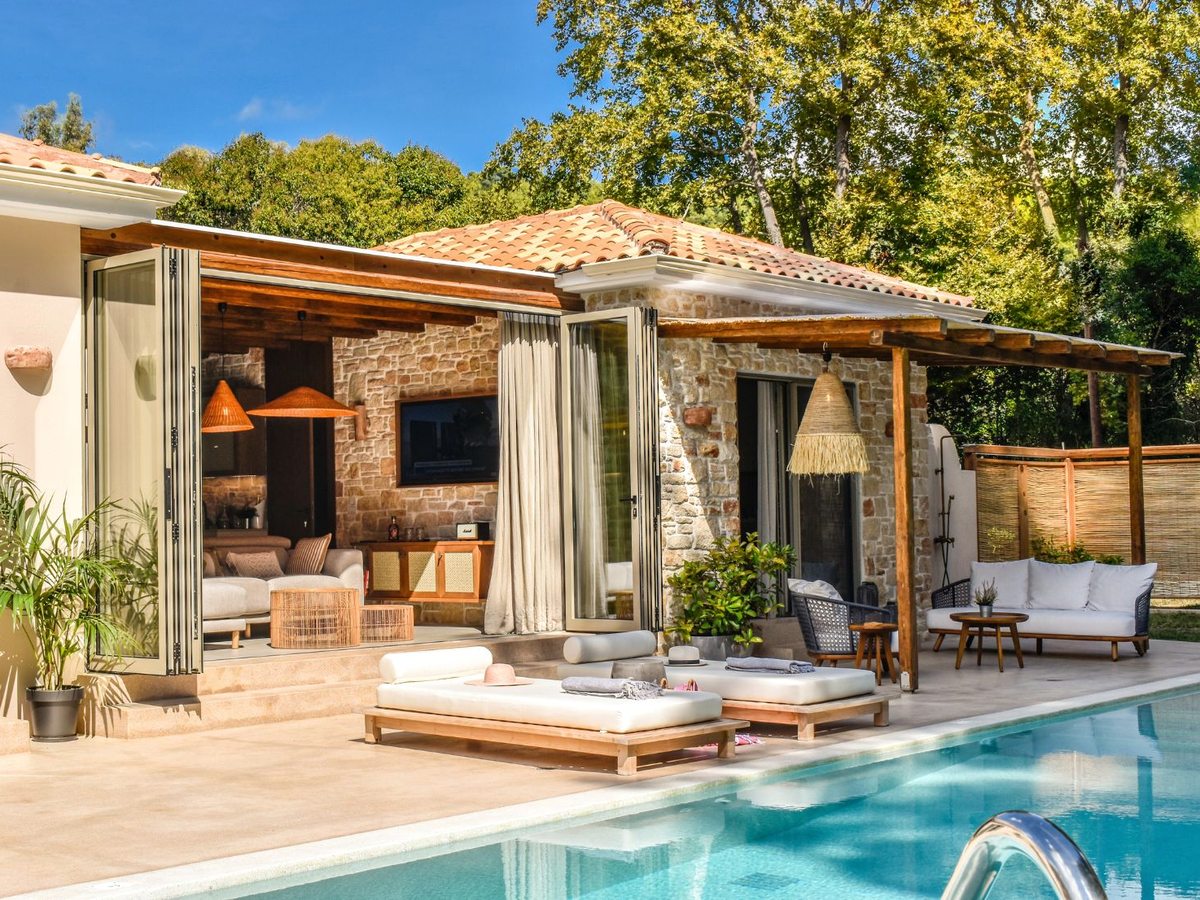 Stone-walled single-storey villa with folding glass doors open onto a terrace, wooden daybeds and a private blue pool in front, backed by trees.