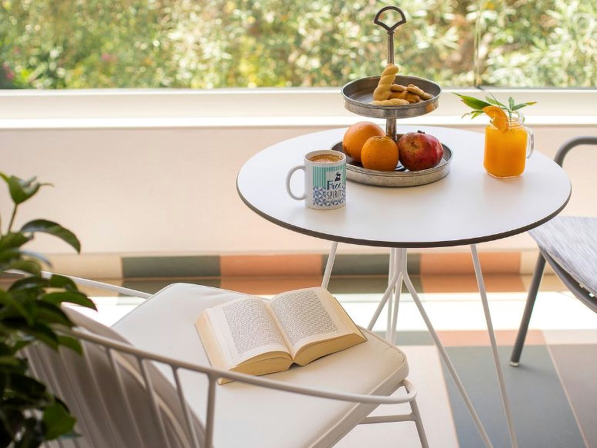 Small round white table on a sunlit balcony set with a tiered stand of Greek bread rings, a tray of oranges and pomegranate, a jar of orange juice and a mug of coffee, with an open book on a cushioned chair.