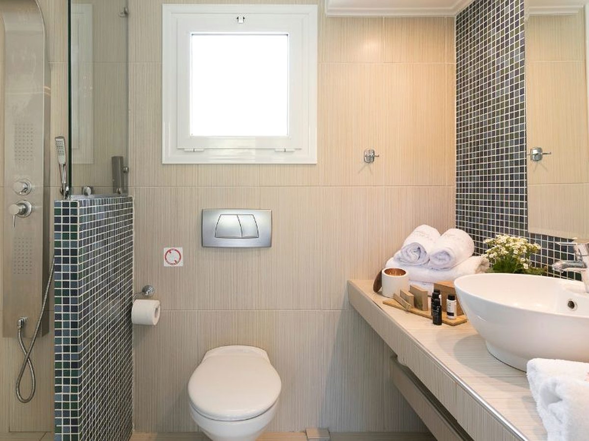 Bathroom with a glass-screened walk-in shower, mosaic tile bands, a wall-hung toilet, a white basin on a pale counter, rolled towels and toiletries.