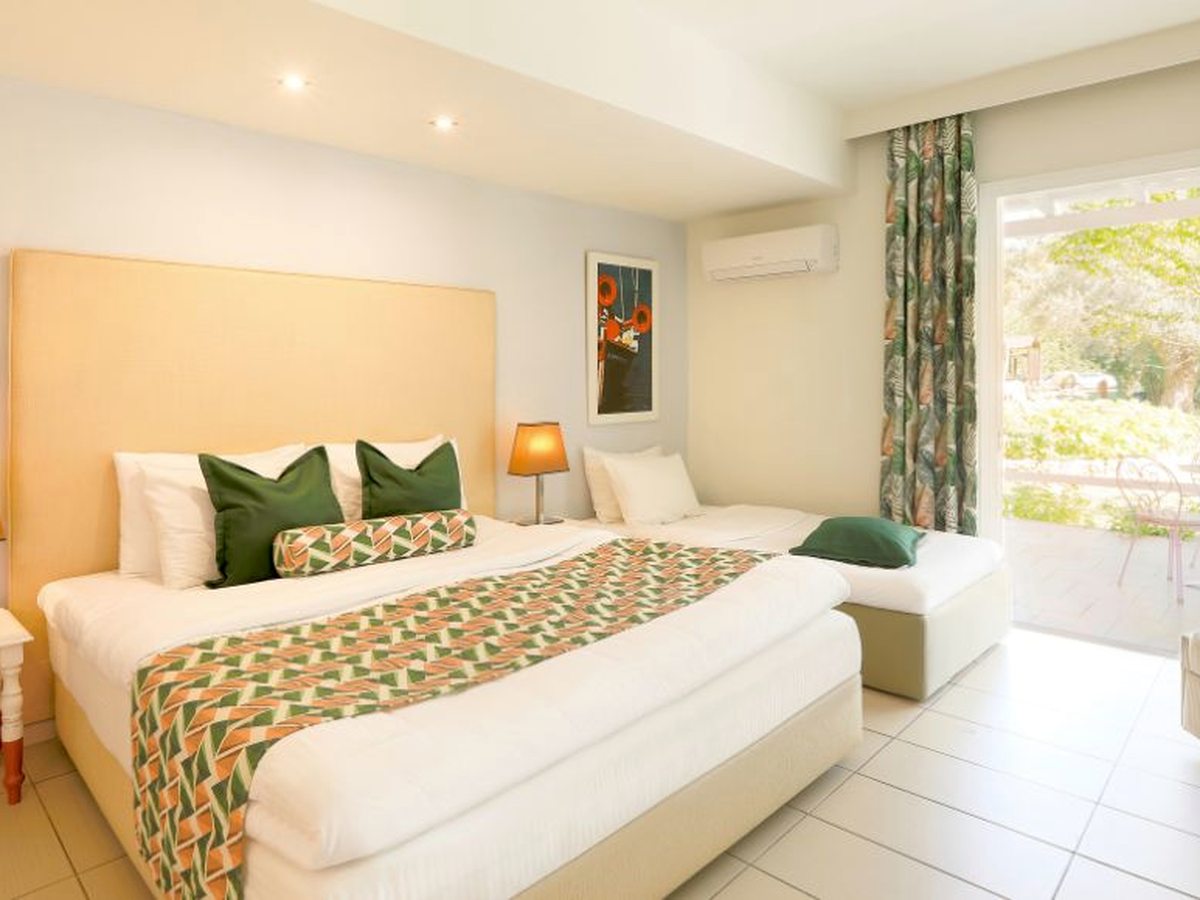 Bright bedroom with a double bed and a single bed side by side, cream headboards, green cushions and a patterned runner, and sliding glass doors onto a terrace with garden beyond.