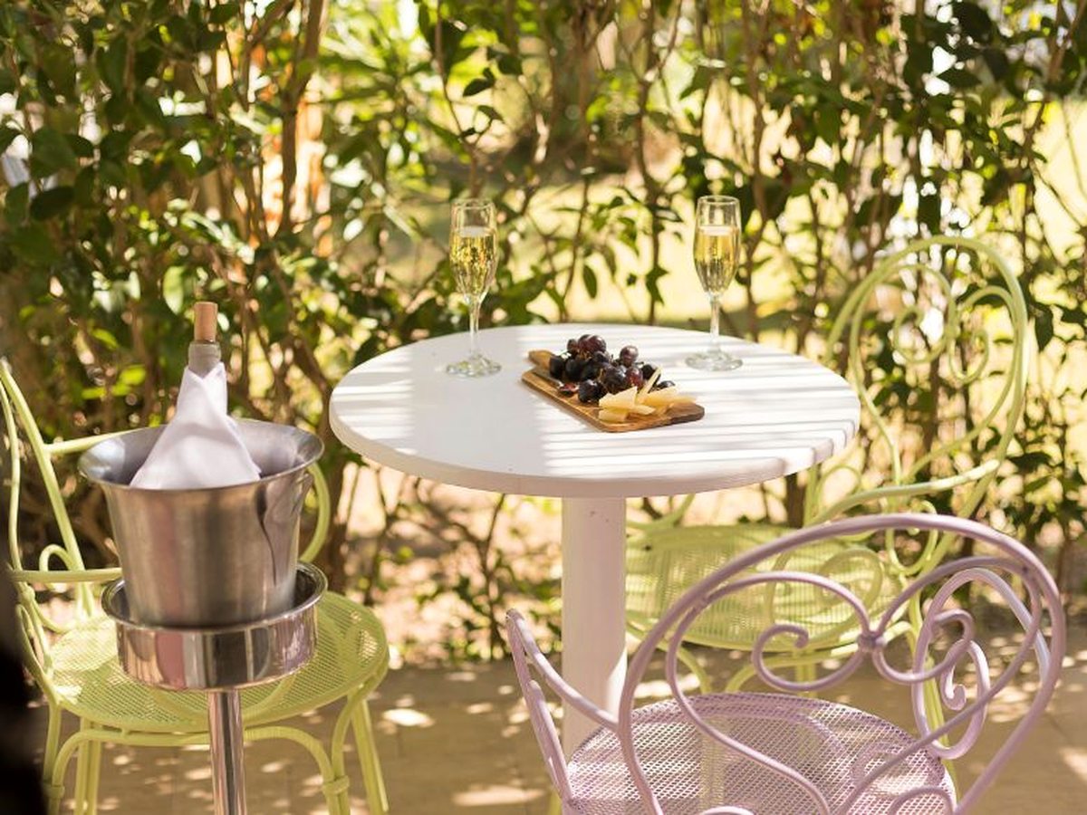 Round white garden table under leafy trees set with two glasses of sparkling wine and a wooden board of black grapes and cheese, with a bottle in a silver ice bucket beside pale green and lilac metal chairs.