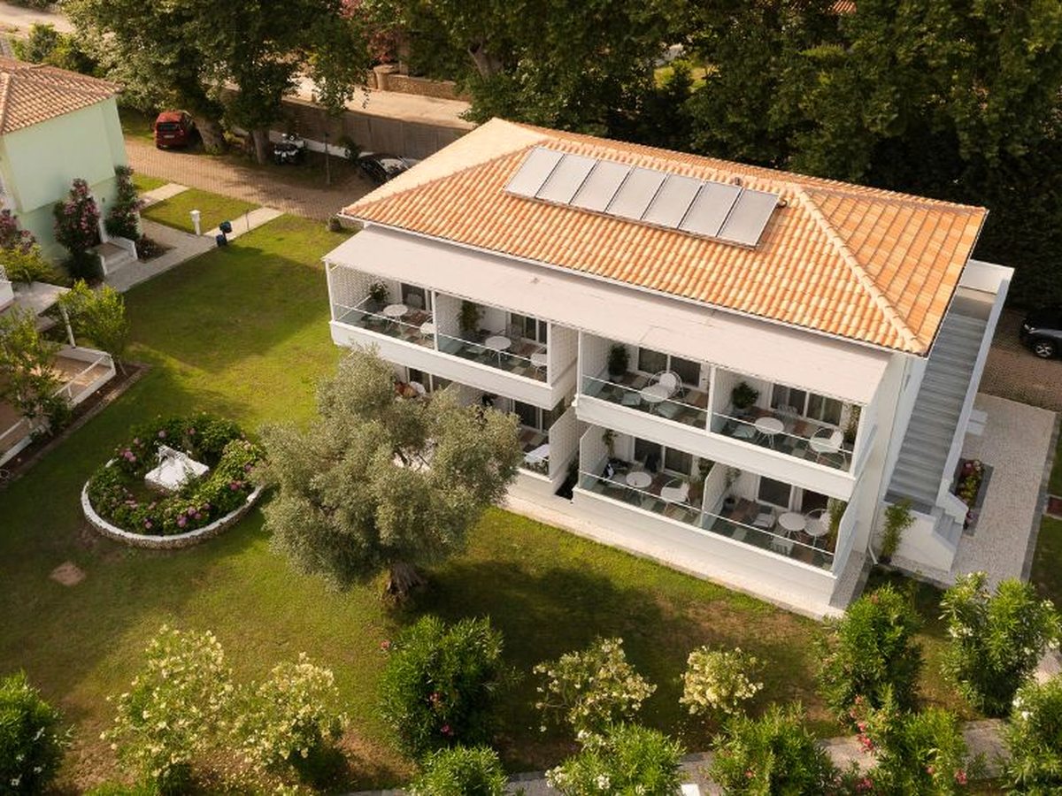 Aerial view of a two-storey white hotel building with a terracotta roof and balconies, surrounded by lawns, an olive tree, a circular flower bed and paths.