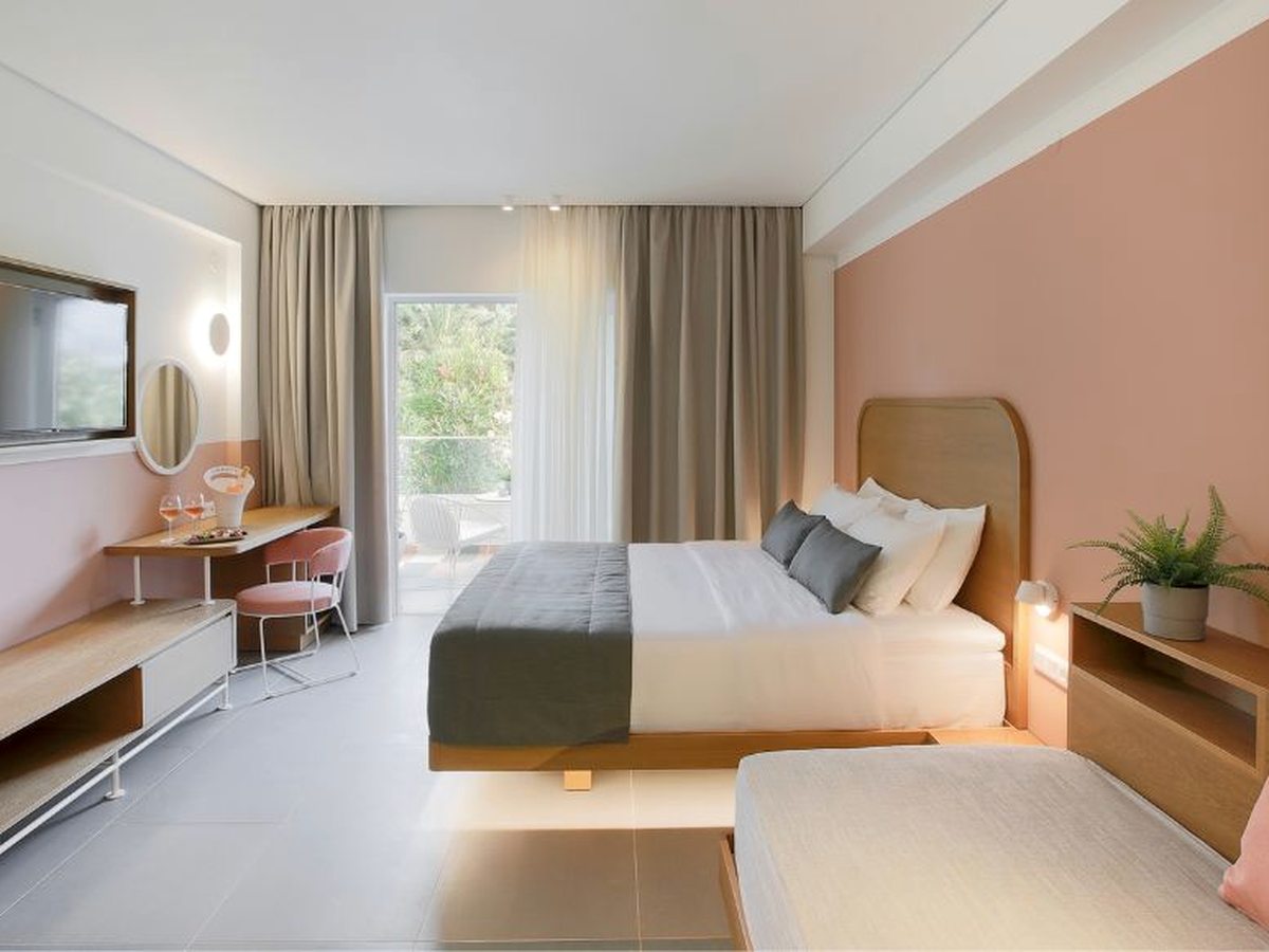 Modern bedroom with a pale pink feature wall, a double bed with a curved wooden headboard, a low sofa bed in the foreground, a desk with a pink chair and glass doors to a balcony.