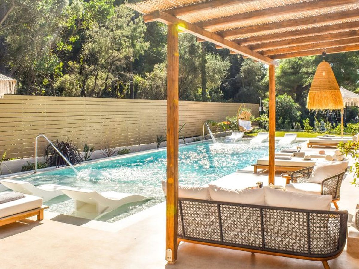 Timber pergola over a poolside lounge with woven sofas and cushioned daybeds, a rattan pendant light, and the pool alongside with white loungers standing in the water.