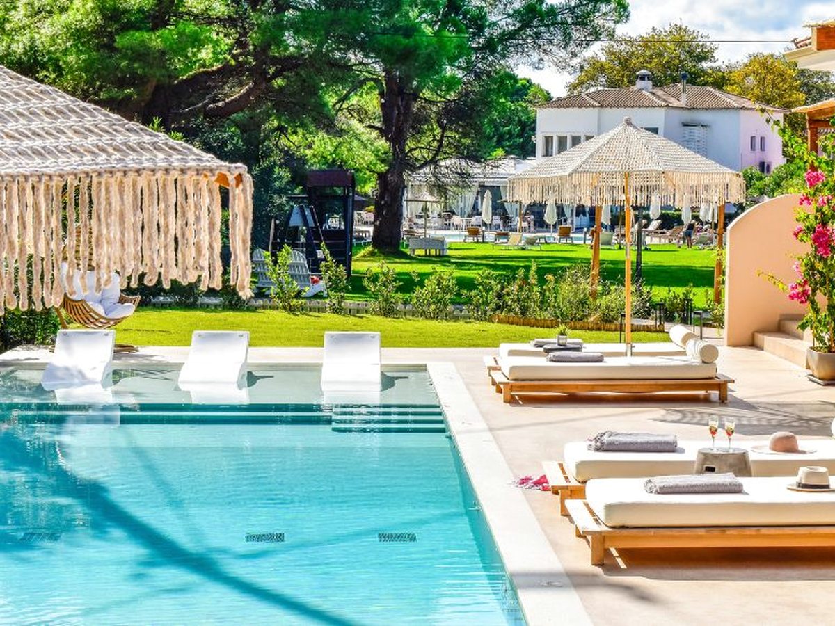 Rectangular blue pool with white loungers standing in the shallow end, wooden daybeds and fringed parasols along the terrace, and a lawn with pine trees and white and pink hotel buildings behind.