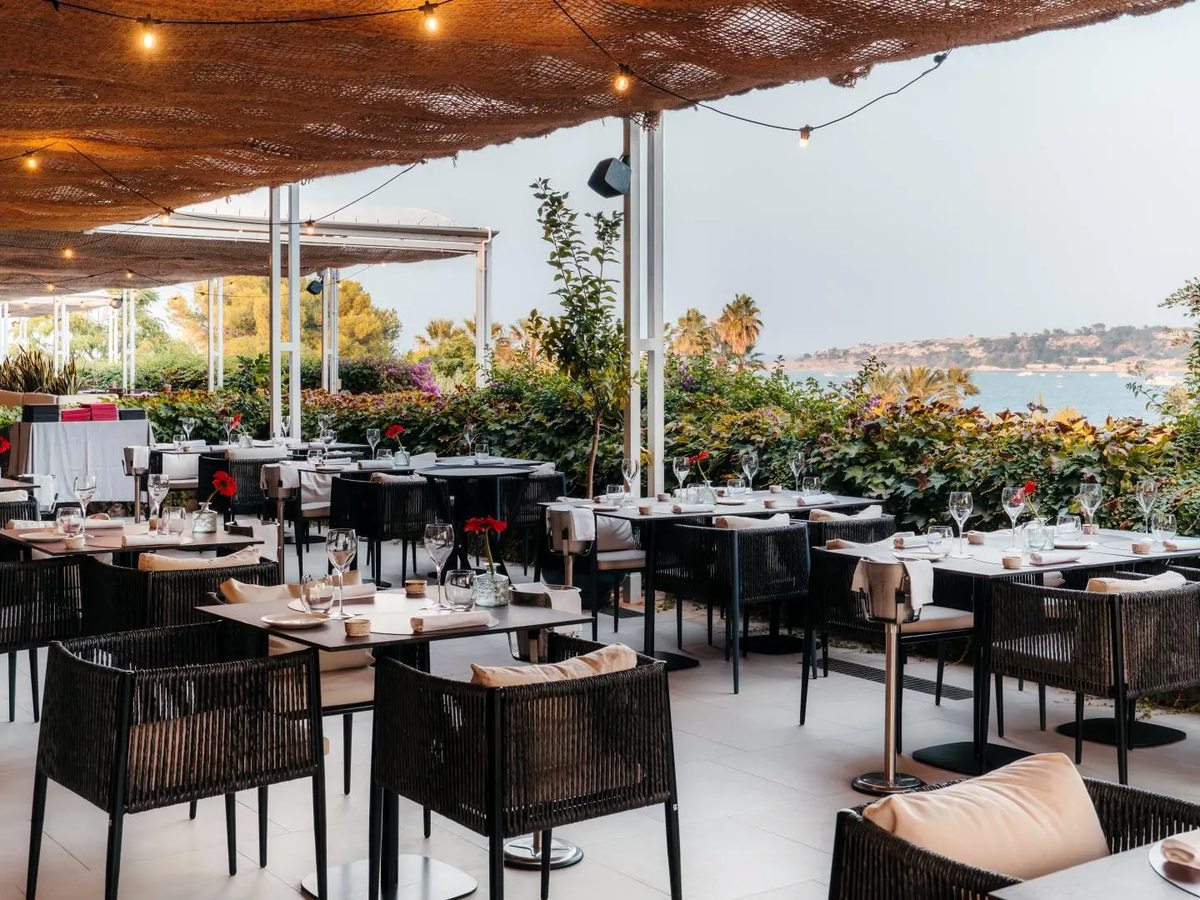 An open-air restaurant terrace under a woven rush canopy strung with festoon lights, with tables laid in white linen and black rope chairs, and the sea and a low headland beyond a planted border.