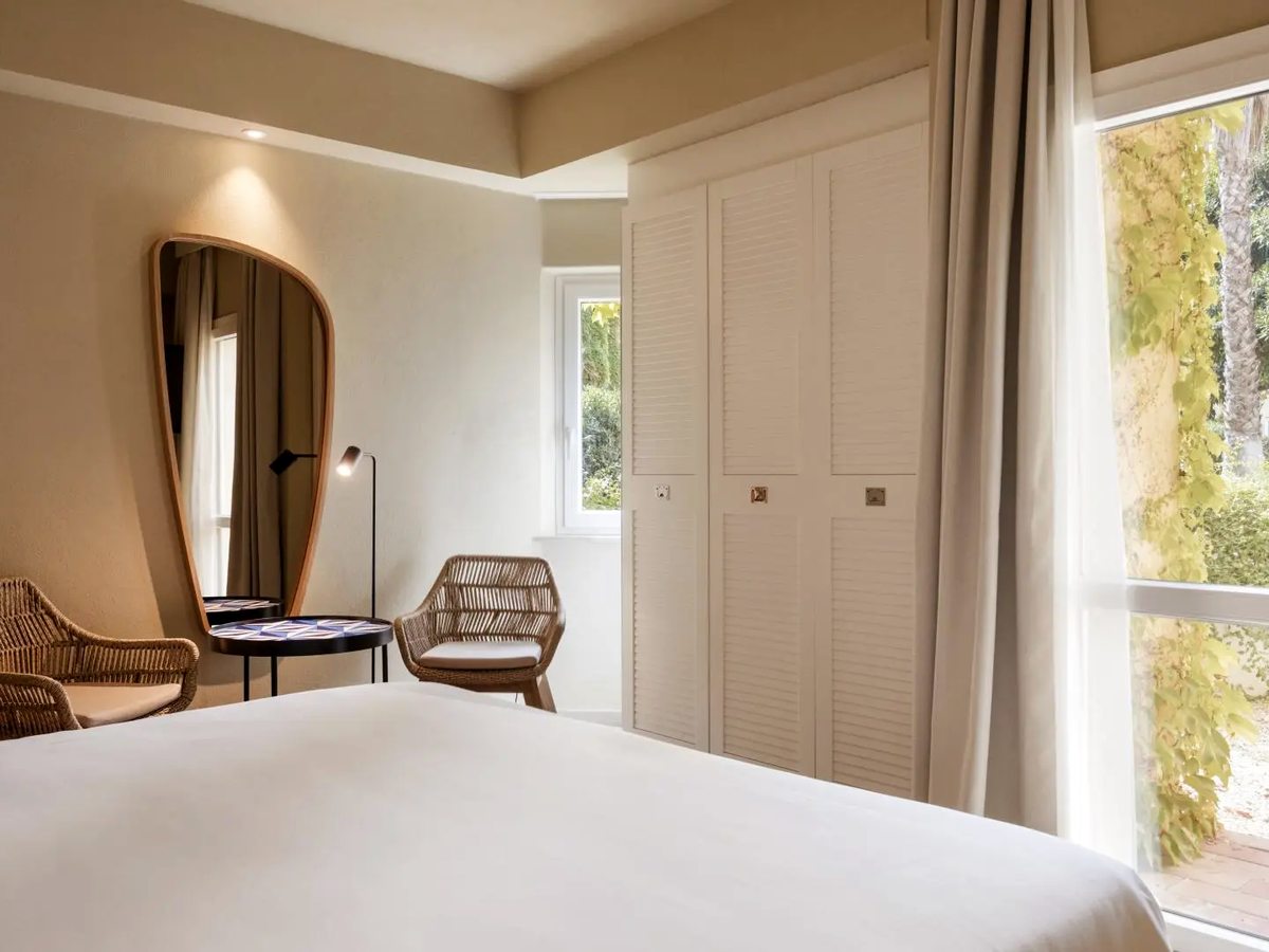A guest room with a white-linen double bed, two wicker armchairs around a small patterned table, a tall shaped mirror and louvred wardrobe doors, with a window onto sunlit greenery.