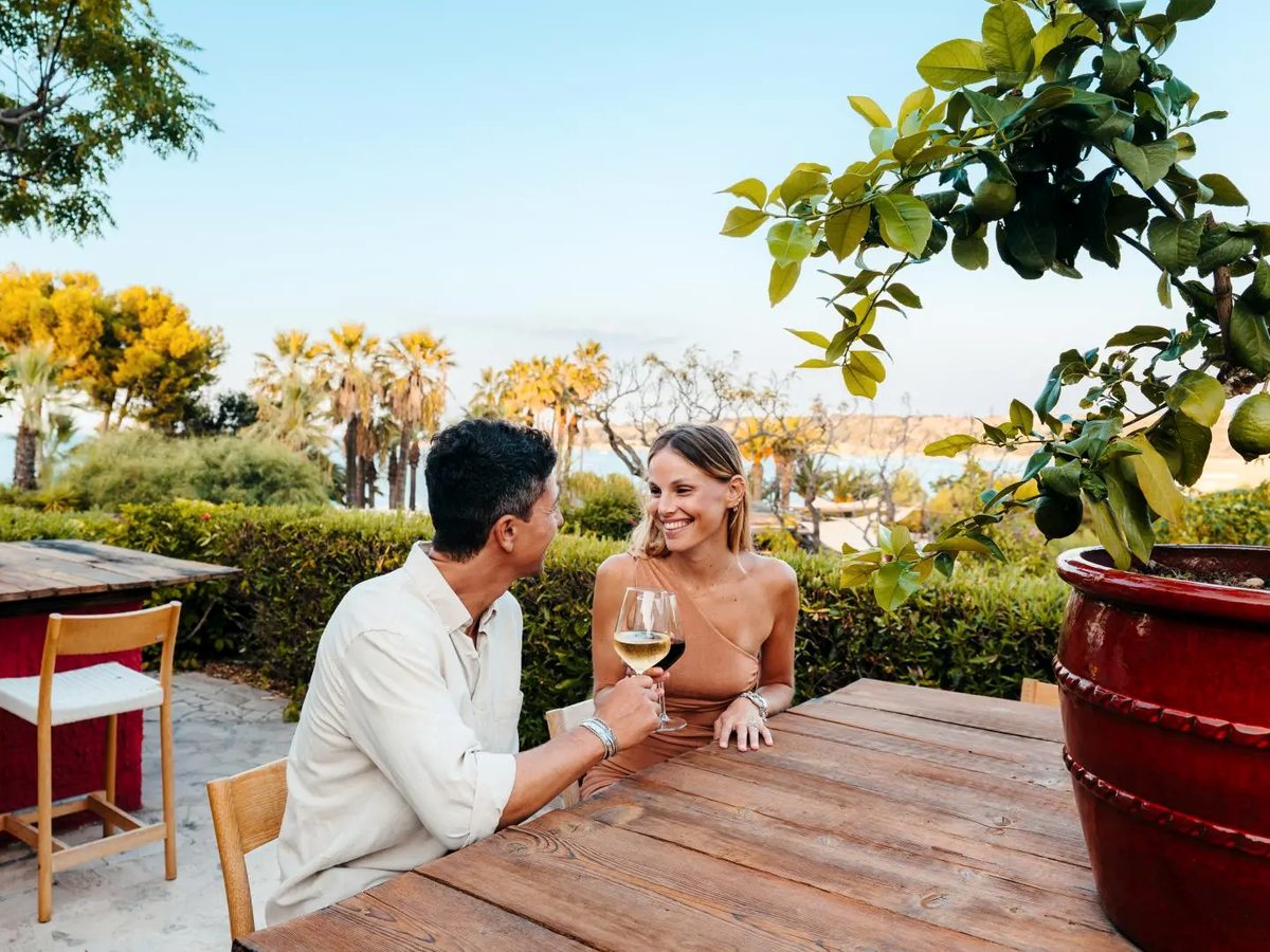 A couple sitting with glasses of white wine at a rustic wooden table on a garden terrace, beside a large red glazed planter holding a lemon tree, with palms and the sea behind.