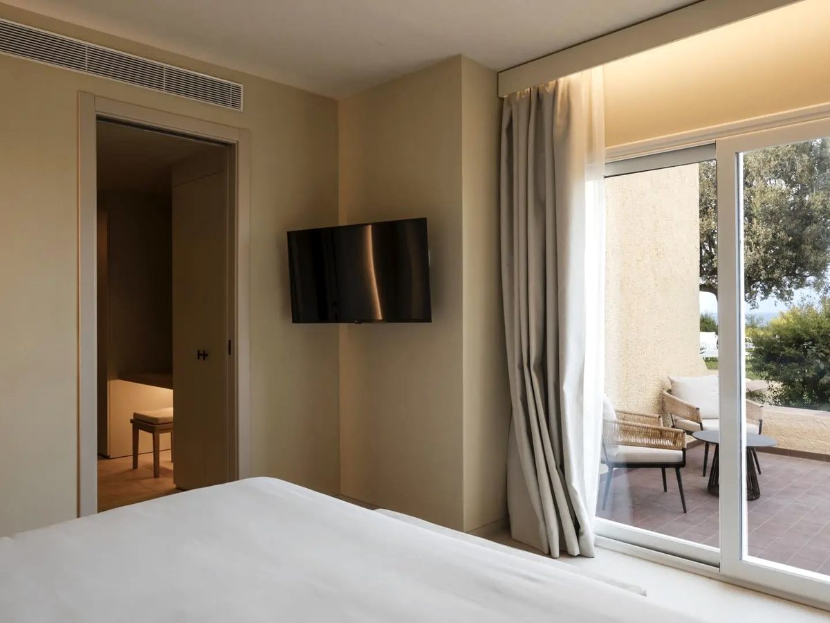A double room with a wall-mounted television and sliding glass doors onto a tiled terrace set with woven armchairs and a round table, with trees and a strip of sea beyond.