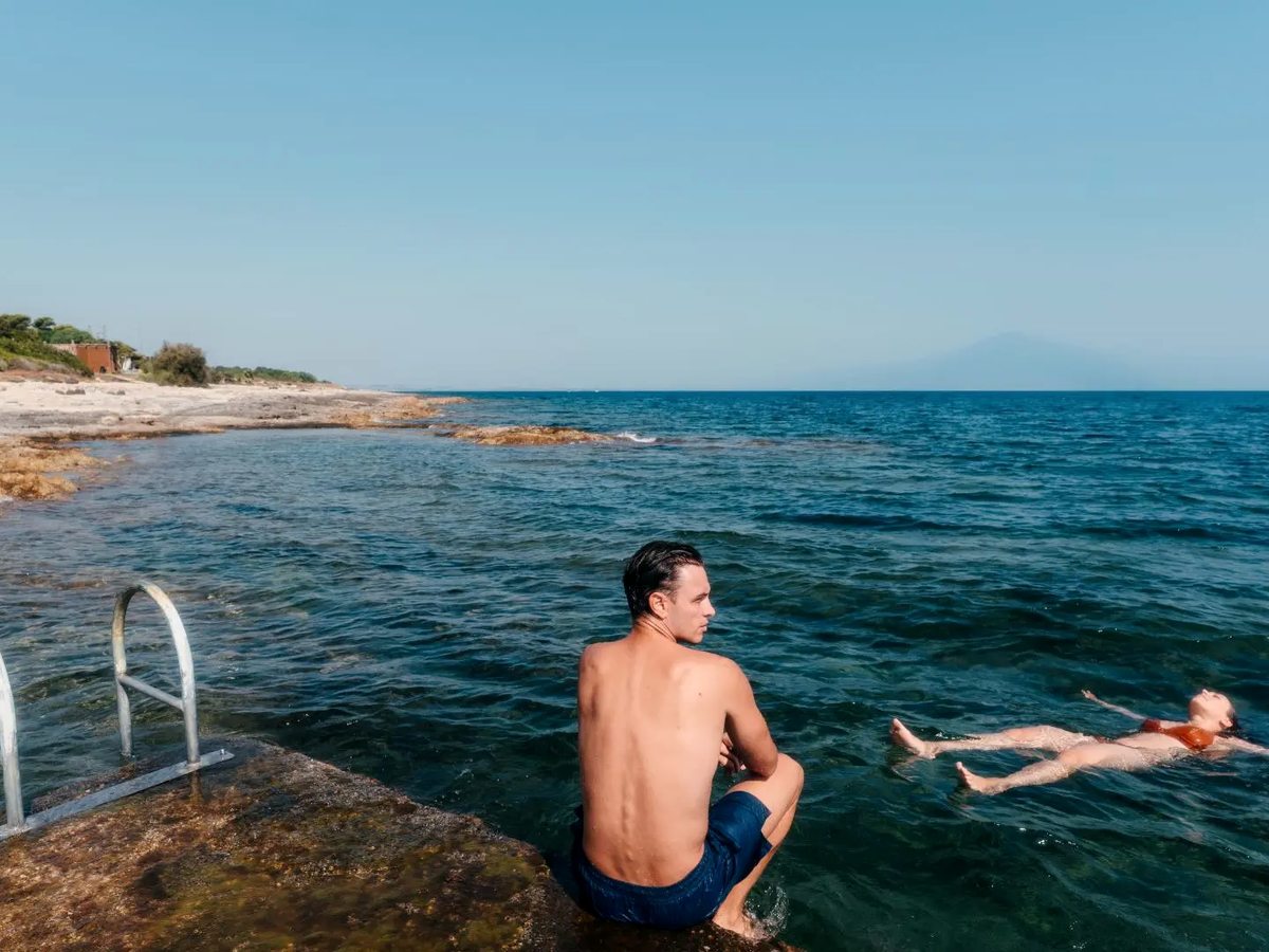 A man sitting on a flat rock beside a metal swimming ladder as a woman floats on her back in clear blue water, with a low rocky shoreline stretching away behind.