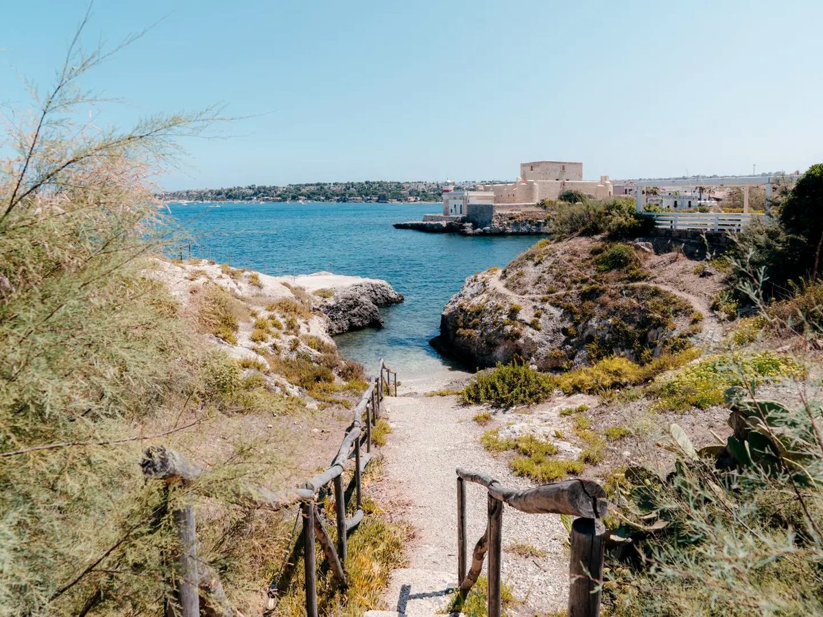 A gravel path with rustic timber handrails running down between rocks and scrub to a small inlet, with a stone fort and a small lighthouse on the far shore under a clear blue sky.