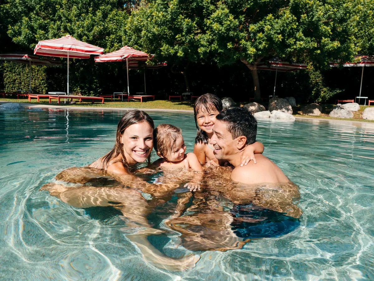Two adults and two young children smiling in the shallow water of a pool edged with boulders, with red-and-white striped parasols and trees behind.