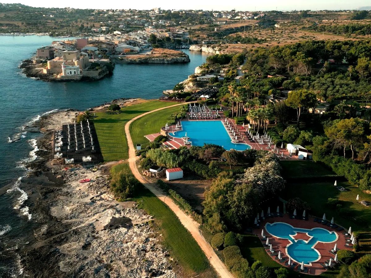 Aerial view of a coastal resort on a wooded headland, with a large rectangular pool and a clover-shaped pool set in lawns above a rocky shoreline, and a village and castle on the headland opposite.