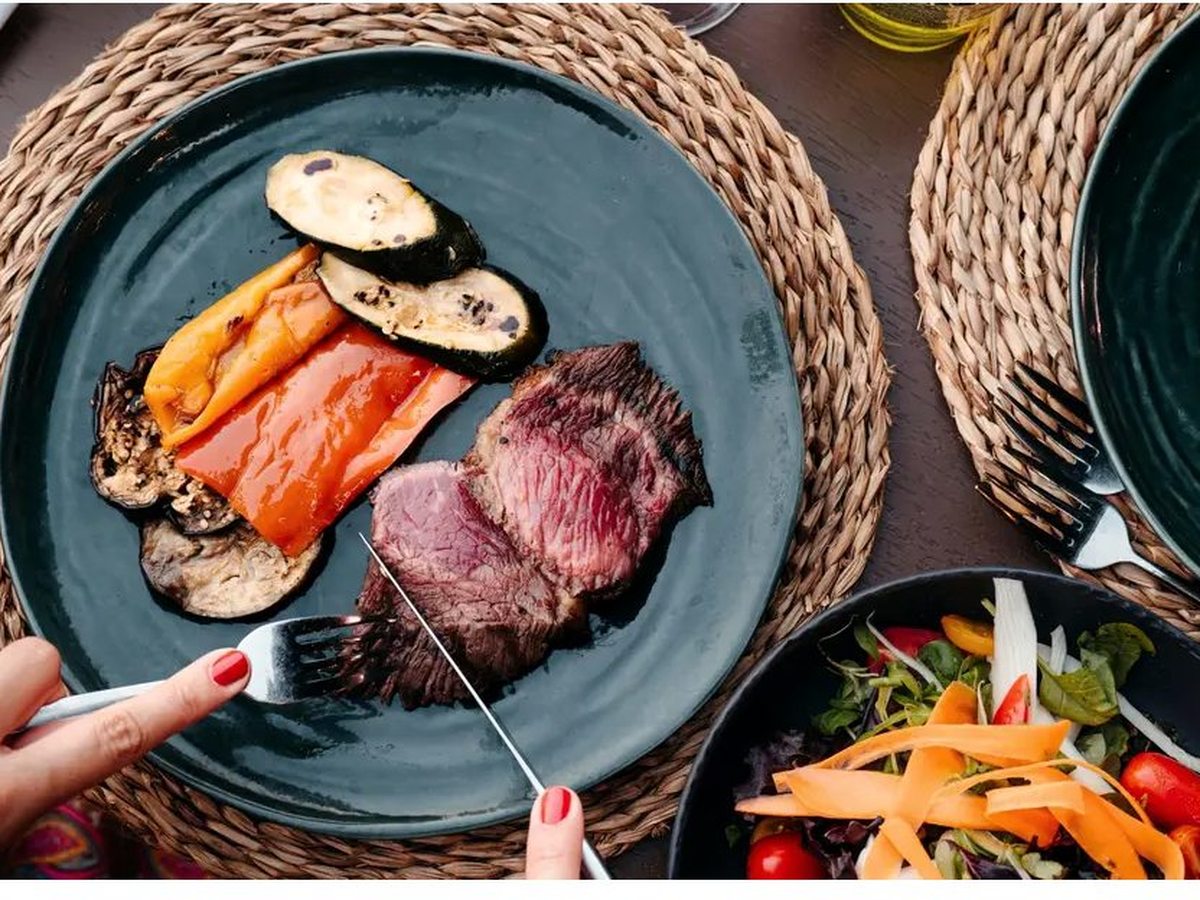 A dark ceramic plate of sliced medium-rare steak with grilled courgette, peppers and aubergine, with a bowl of mixed salad alongside on a woven mat.