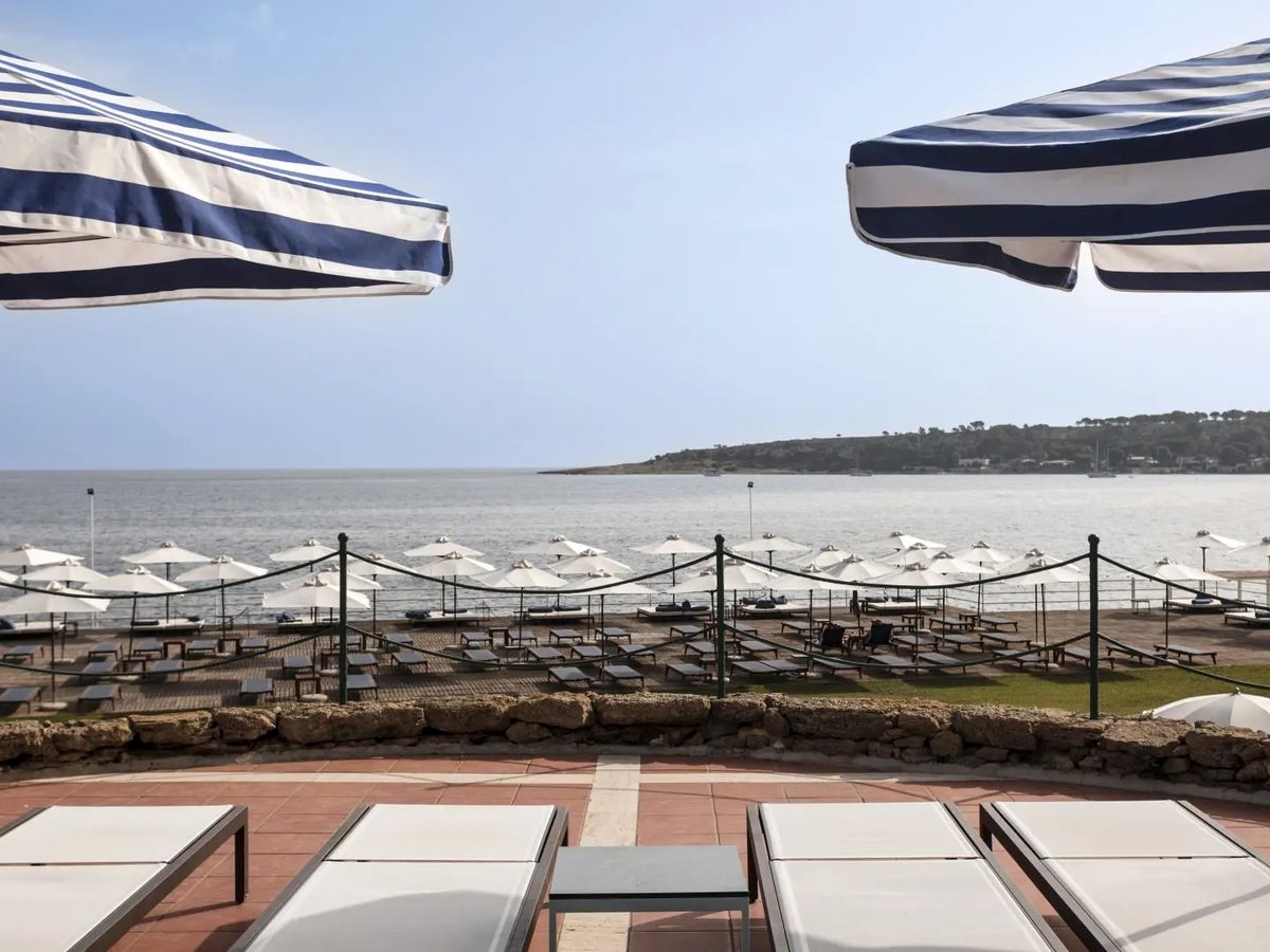 Sun loungers on a terracotta-tiled terrace behind a low stone wall, with rows of white parasols and further loungers on the level below beside the sea.