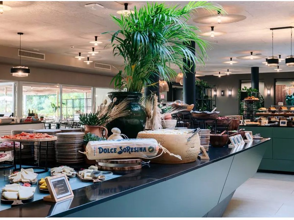 A long dark-green buffet island laid with whole cheeses, stacked plates and platters of cured meats, under pendant lights in a large open dining room with a tall potted palm.