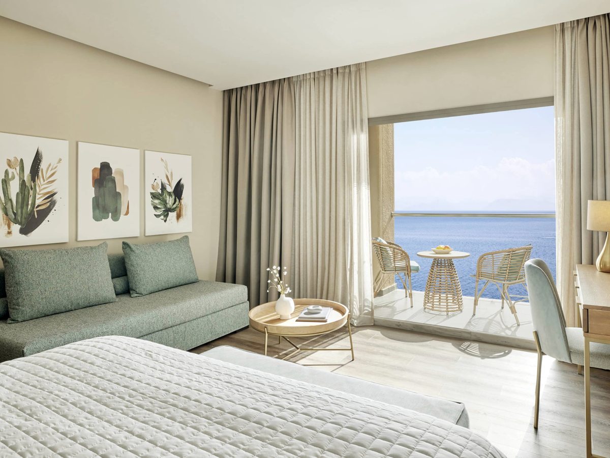 A double bed with a green sofa bed alongside, a desk and chair, and glass doors onto a balcony with two chairs and a table above the sea.