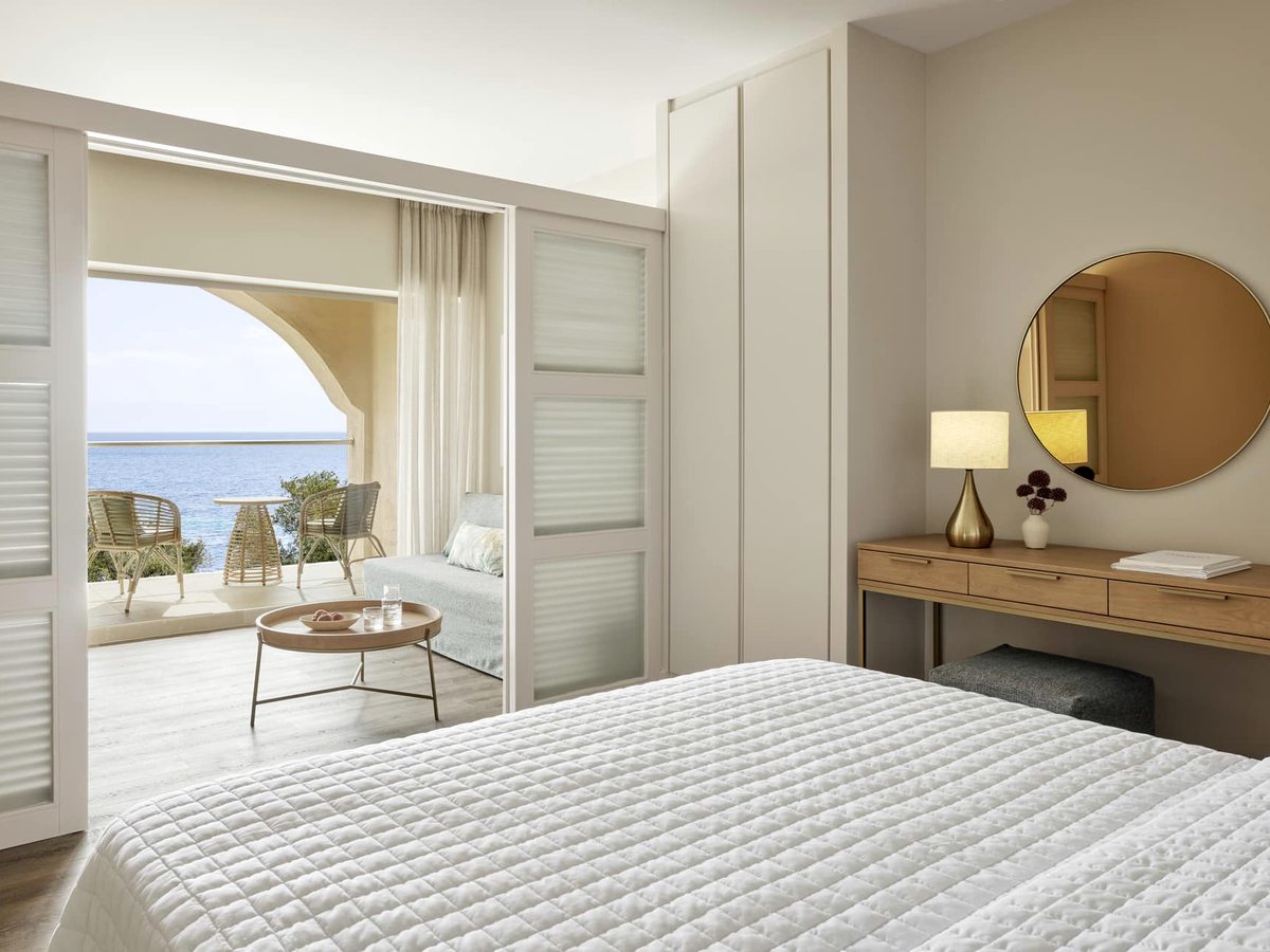 A quilted double bed beside a dressing table and round mirror, with sliding frosted doors open to a sitting area and an arched balcony above the sea.