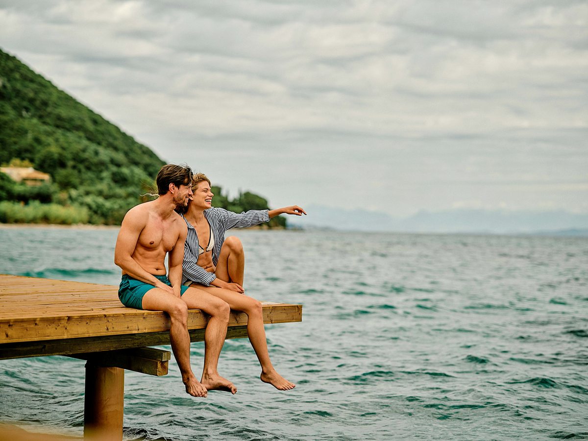 A couple sitting at the end of a wooden jetty with their feet over the water, a wooded headland behind and mainland mountains on the horizon.