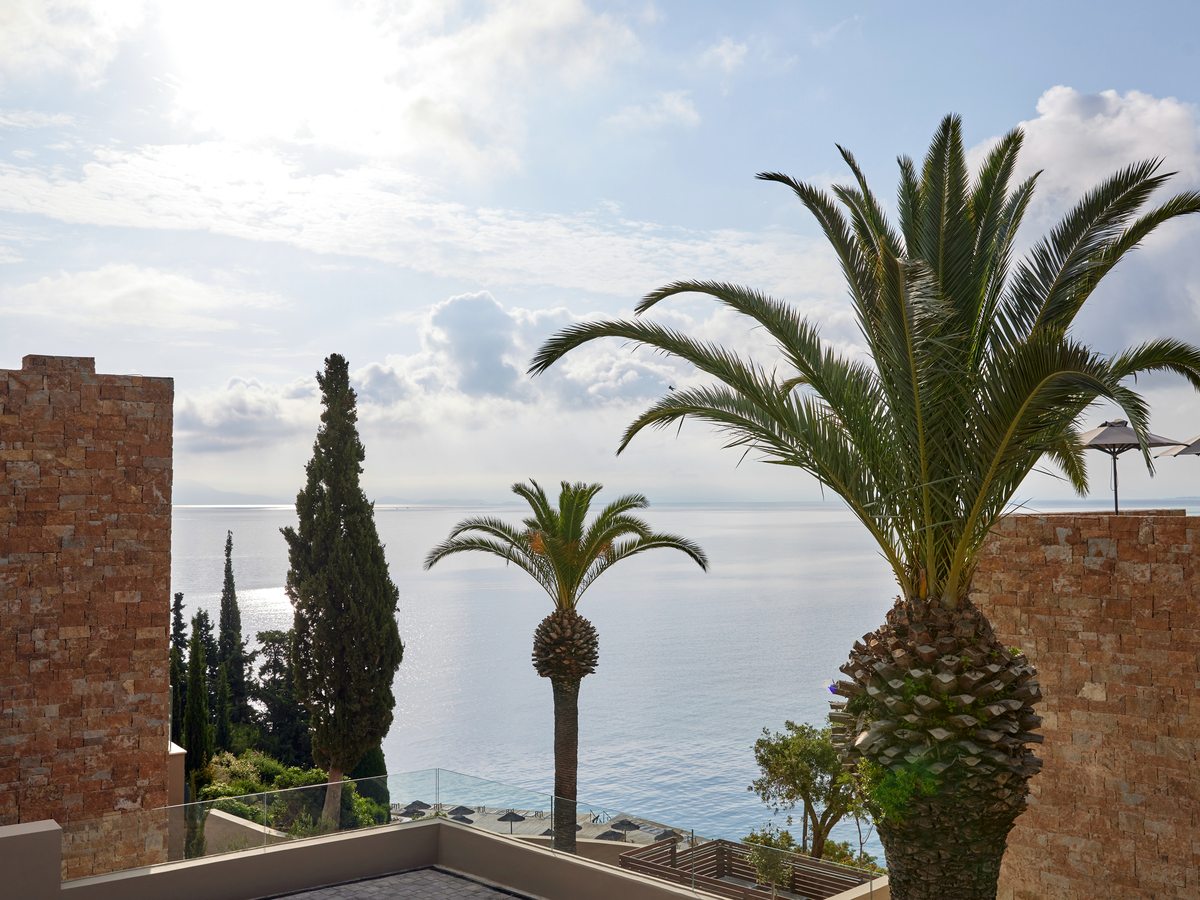 Palms and a cypress framing a stone terrace wall above a calm blue sea, with the mountains of mainland Greece low on the horizon.