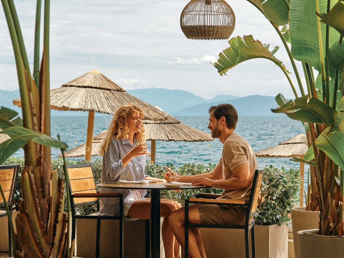 Two people at a small table on a covered terrace beside the beach, with thatched parasols, potted plants and the sea and mountains behind.