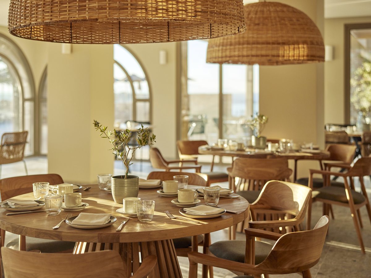 A restaurant interior with round wooden tables laid for a meal, cane-backed chairs, large woven pendant shades and arched windows.