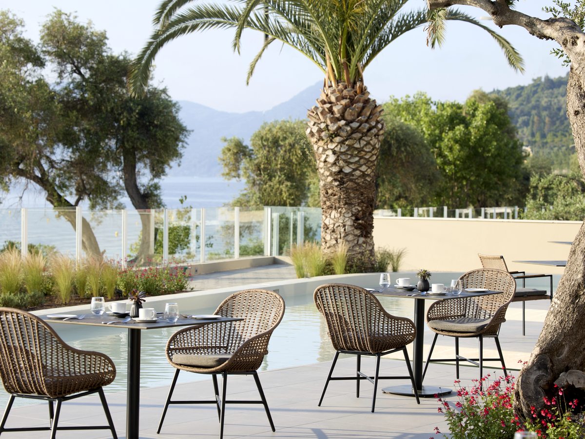 Outdoor tables with woven armchairs on a paved terrace beside a shallow water channel, under palms and olive trees with the bay beyond.