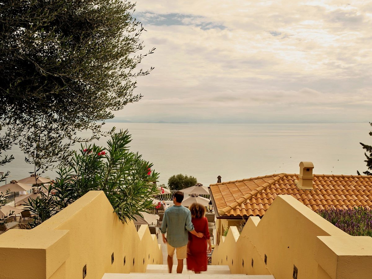 A couple walking down a wide flight of pale stone steps between planted walls, with terracotta roofs, sun parasols and the sea below.