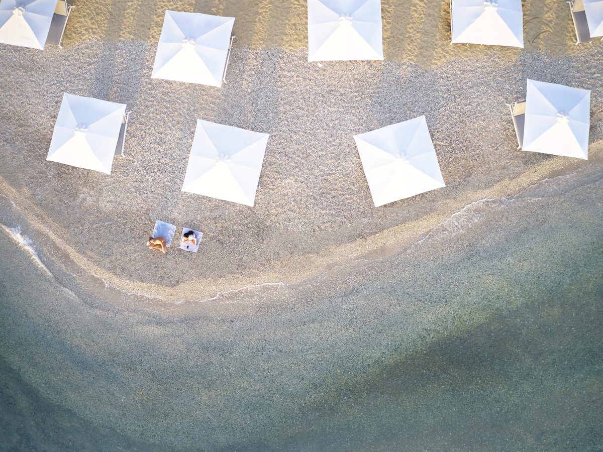 White square parasols and sun loungers set out on grey shingle beside clear shallow water.