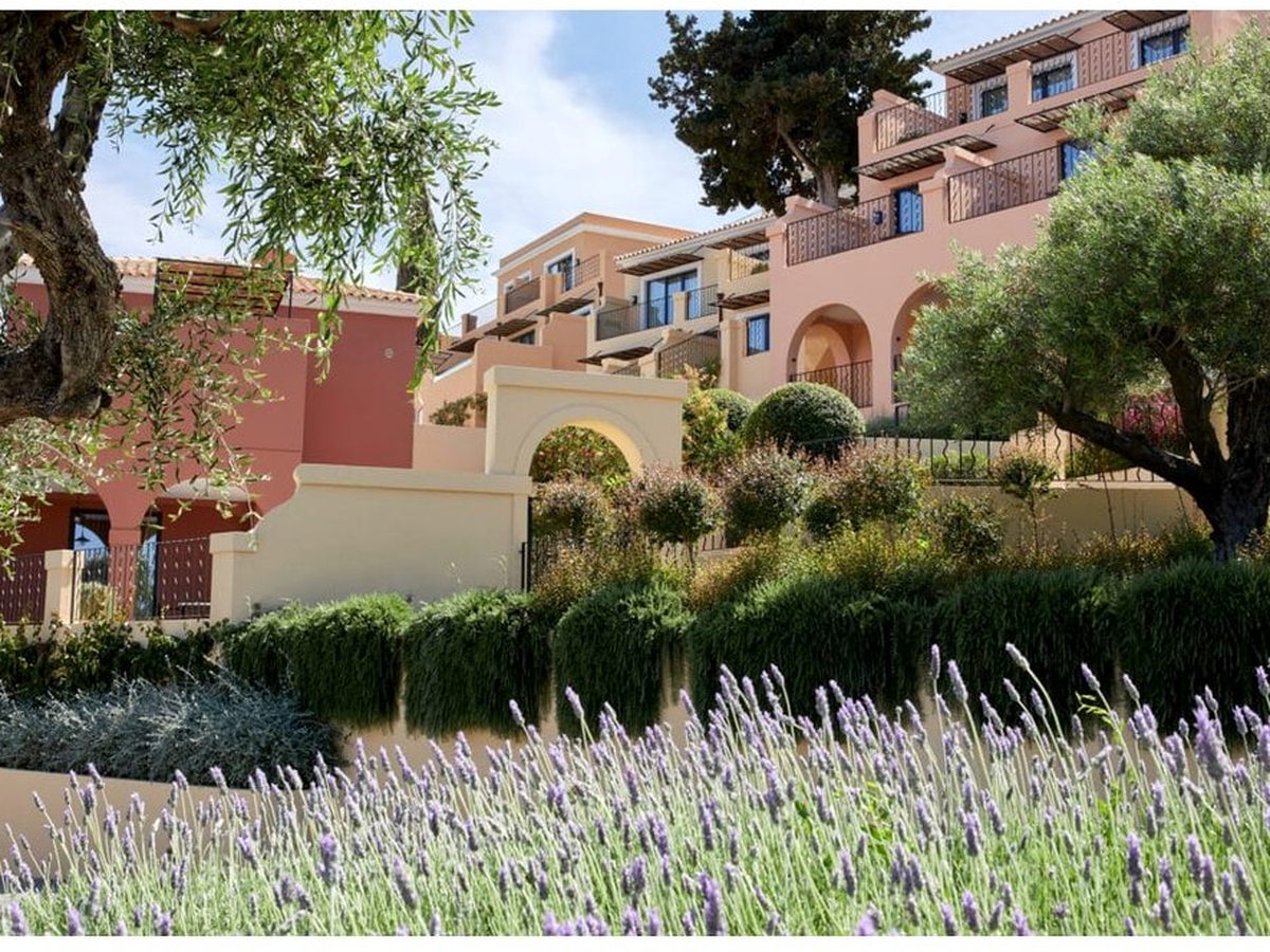 Apricot and terracotta buildings with arched loggias and balconies stepping up a hillside behind olive trees, clipped hedges and flowering lavender.