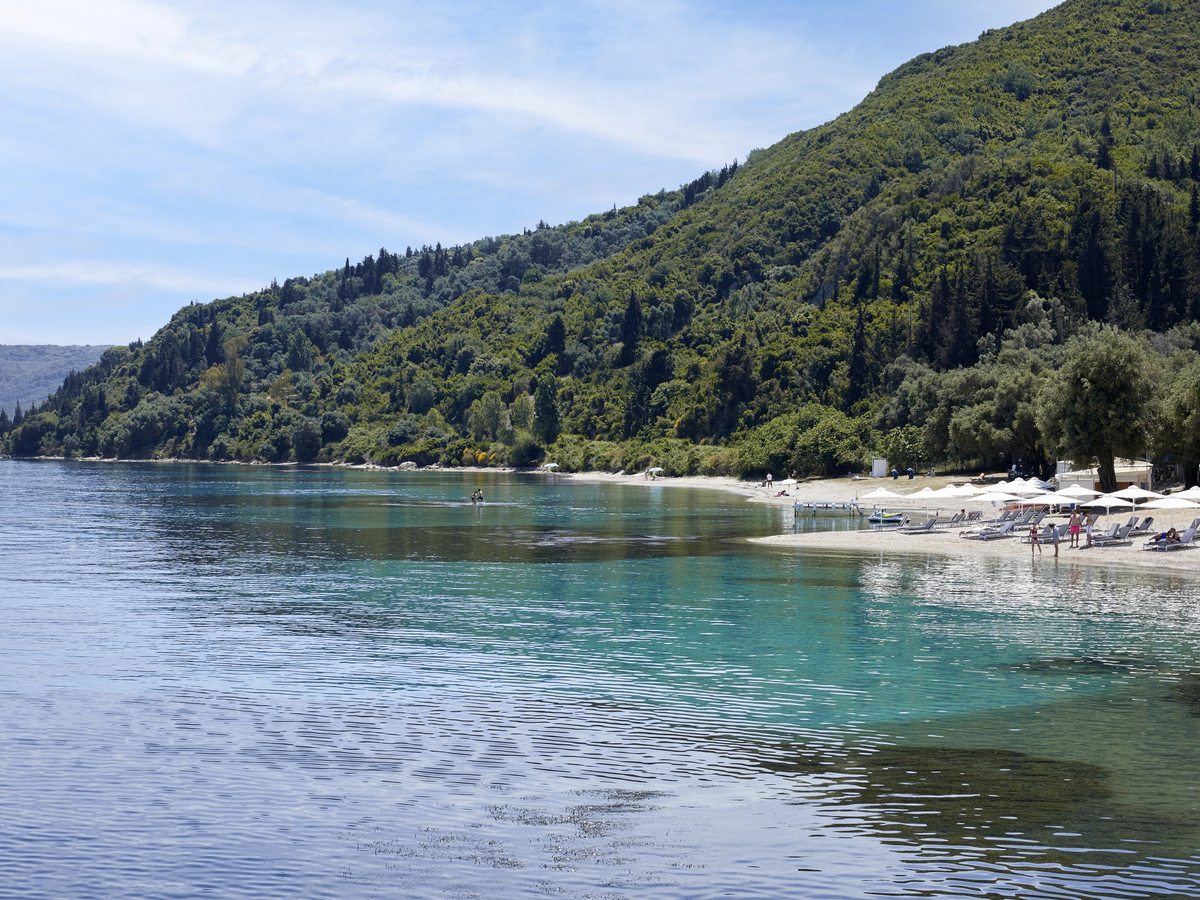 A curving shoreline of pale pebbles with white parasols and loungers at one end, clear green water in the foreground and a wooded hillside behind.