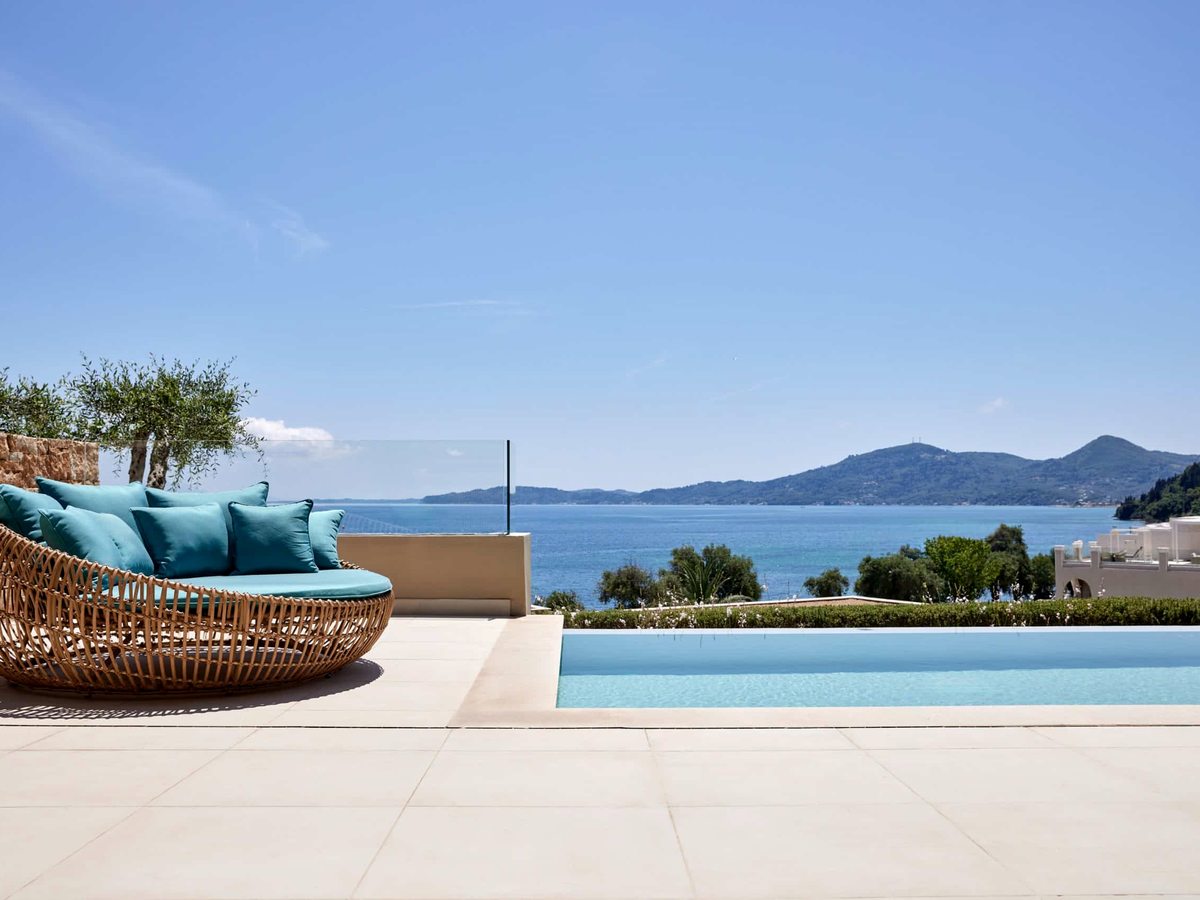 A round rattan daybed with turquoise cushions on a stone terrace beside a small infinity pool, with olive trees and the bay and hills beyond.