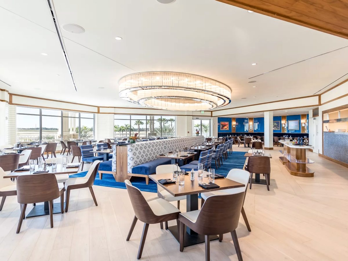 A restaurant dining room with upholstered chairs, curved banquette booths, circular chandeliers and floor-to-ceiling windows.