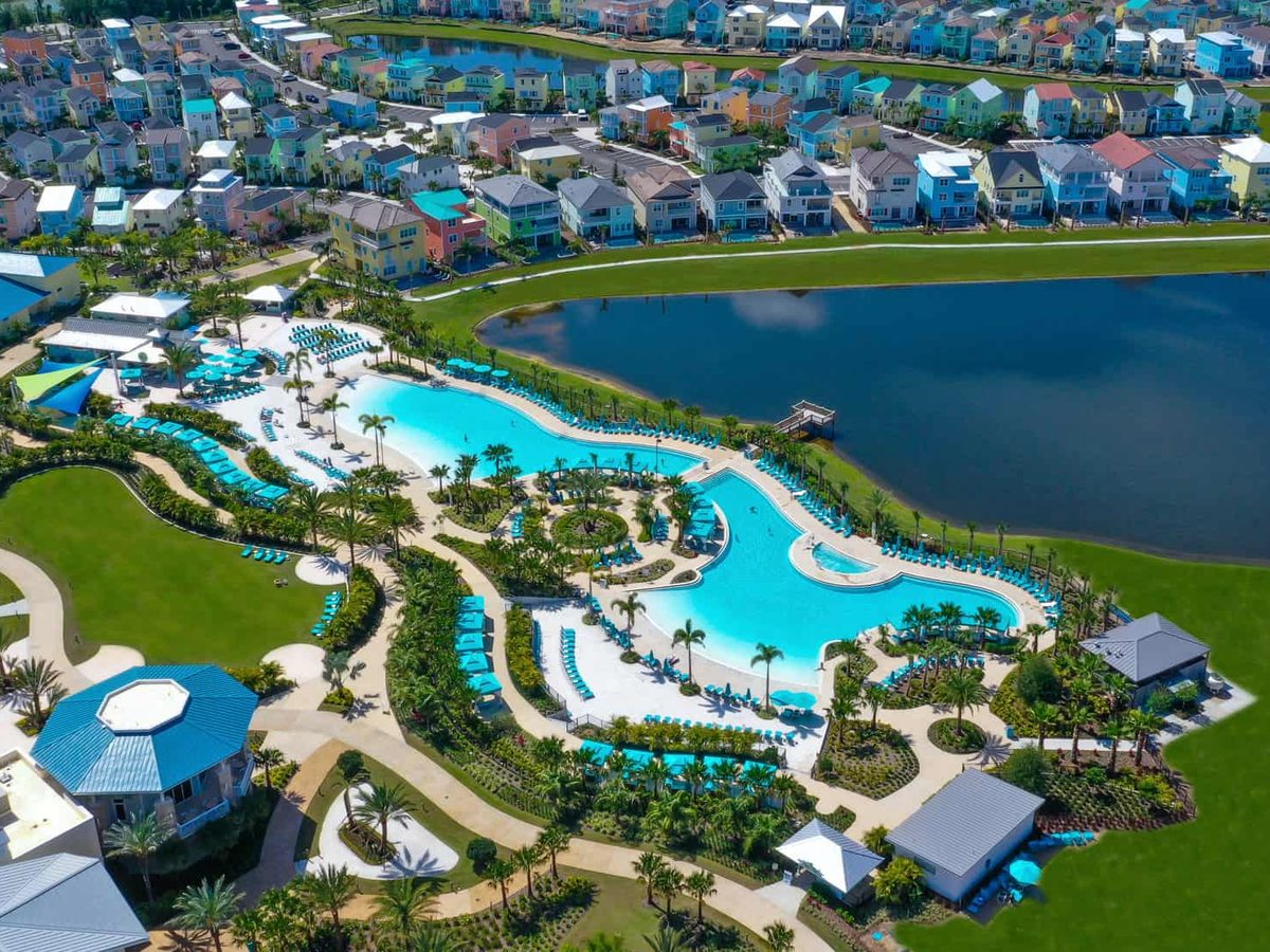 Aerial view of a large freeform turquoise lagoon pool with curving decks and planted islands, beside a lake, pastel resort buildings around it.