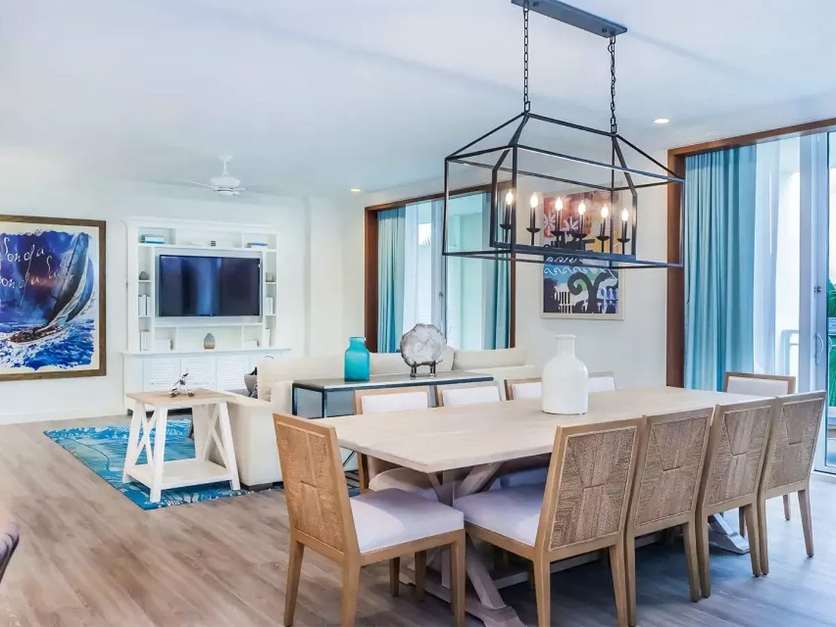 An open-plan living and dining room with white sofas, a wall-mounted TV, a long timber dining table under a rectangular chandelier, wood floor and a ceiling fan.