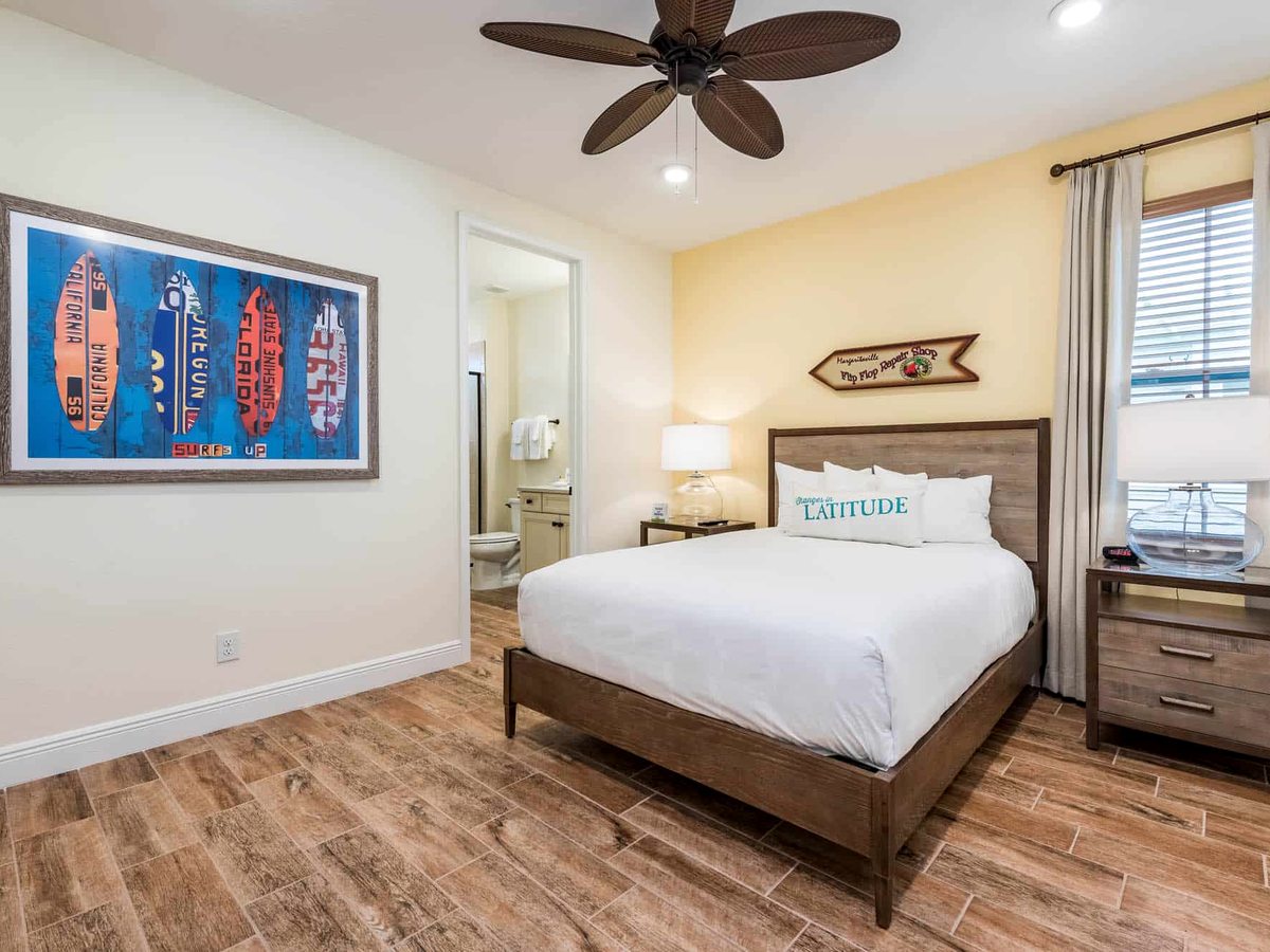 A cottage bedroom with a double bed, a 'Latitude' slogan cushion, bedside lamps, a ceiling fan and surf-themed wall art, an en-suite beyond.