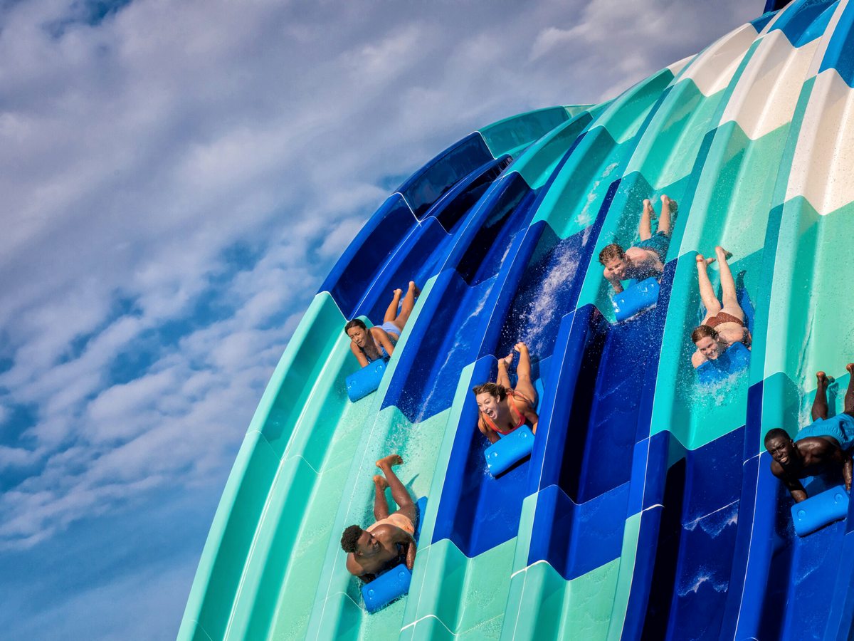 Several parallel racing water slides dropping steeply, with riders going head-first on mats, in turquoise, blue and white.