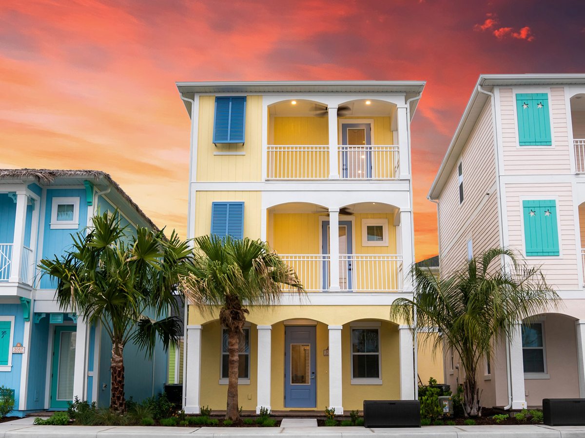 A row of three-storey pastel cottages in yellow, green and teal with balconies and palm trees in front, under a pink and blue evening sky.