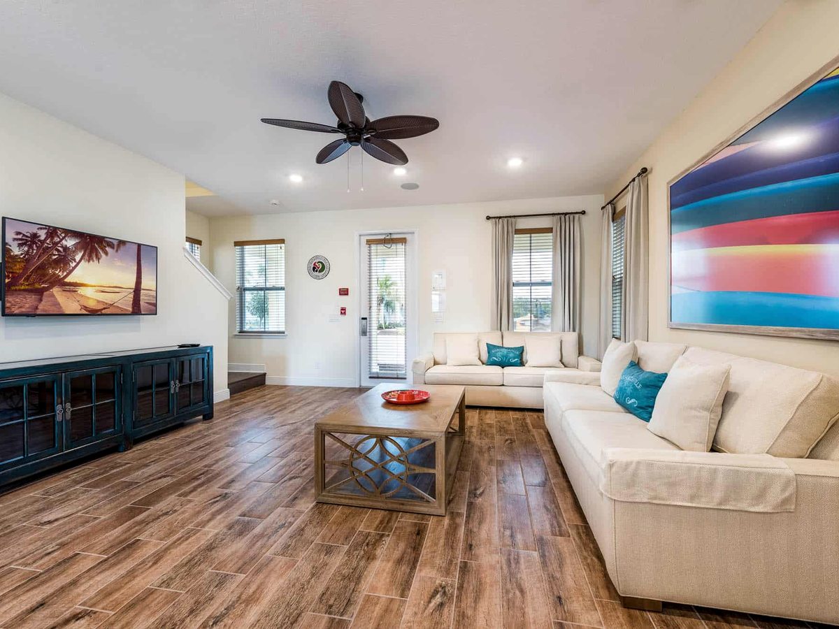 A cottage lounge with a cream sectional sofa, wooden coffee table, wall-mounted TV on a teal console and a vivid sunset painting above, wood floor and ceiling fan.