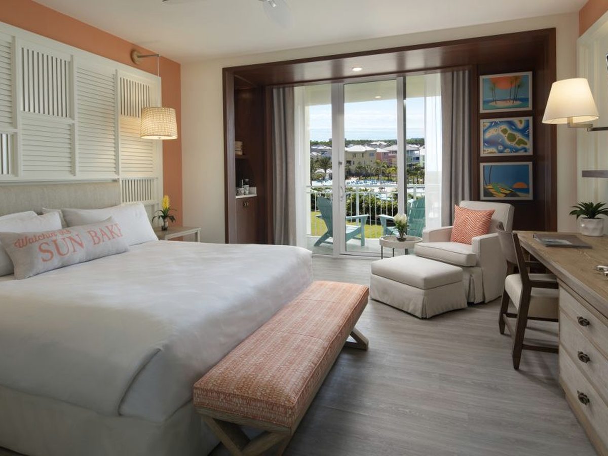 A hotel room with a king bed, a chaise and bench at its foot, a writing desk, wall-mounted TV and glass doors to a balcony over the grounds.