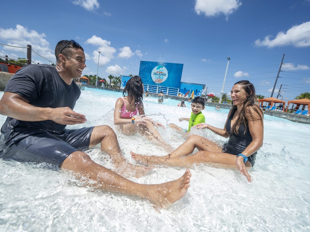 A wave pool with a breaking wave and foaming water, parents and young children standing in the shallow end, water-park buildings behind.