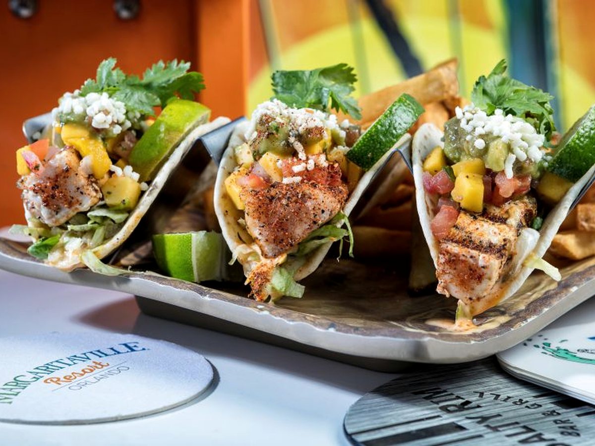Three grilled fish tacos in soft tortillas with shredded greens, fruit salsa, crumbled cheese and lime, with fries, on a metal tray.