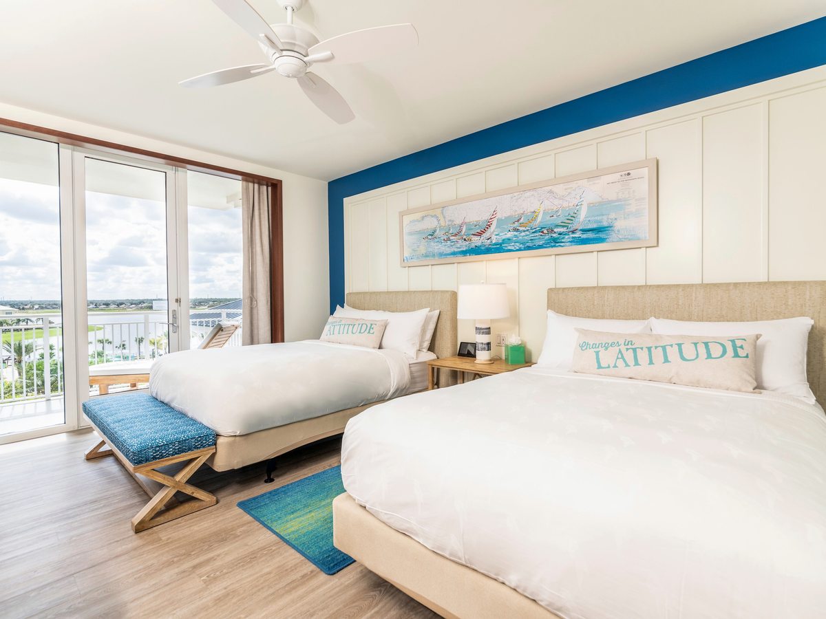 A hotel room with two queen beds under a blue feature wall, matching lamps, a footstool bench and glass doors to a balcony with a pool view.