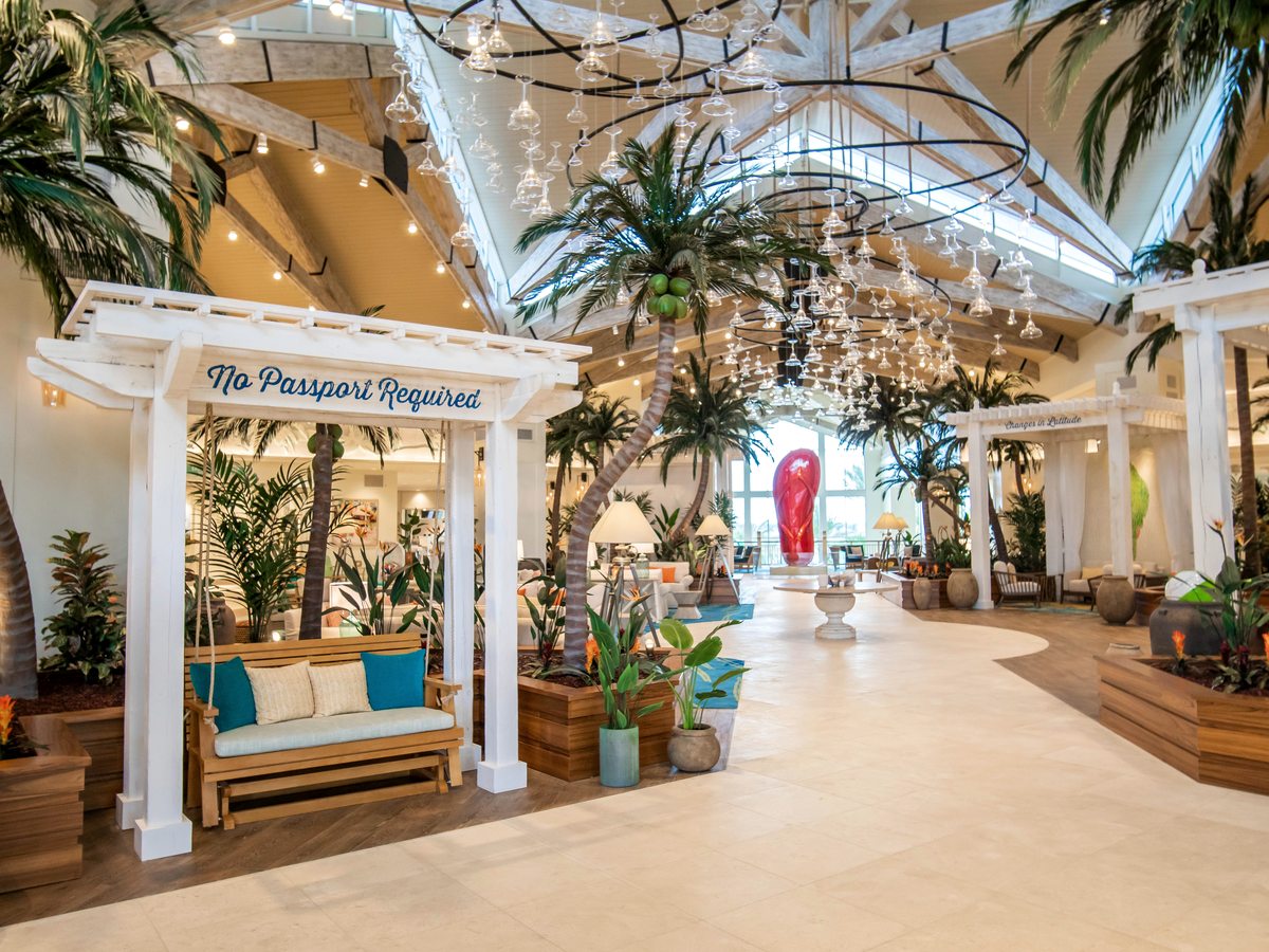 A bright indoor lobby atrium with a white pergola reading 'No Passport Required', tall potted palms, a vaulted timber ceiling and a tiled floor.