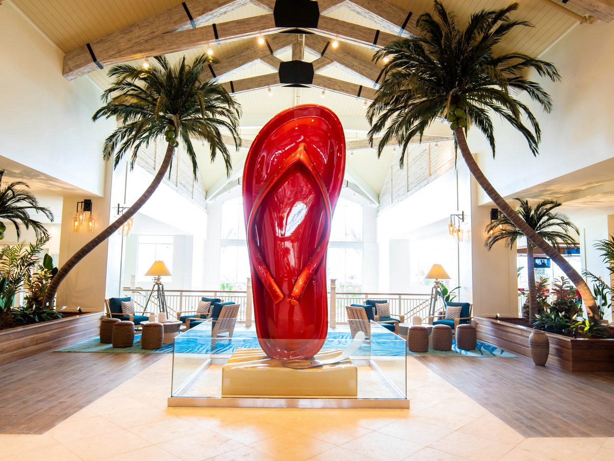A tall red glass sculpture of a flip-flop on a lit pedestal in a lobby with palms, timber roof beams and tall windows.
