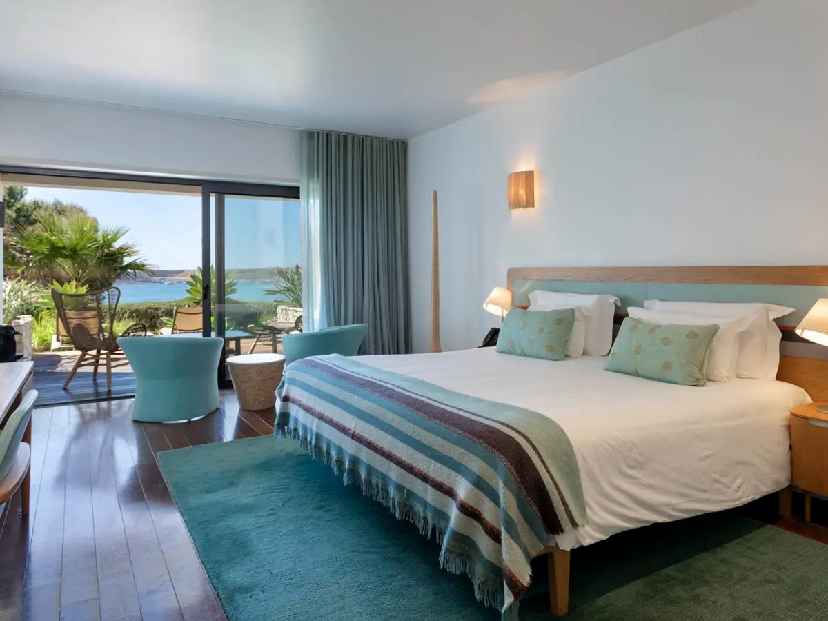 A bedroom with a wooden headboard, white bedding and turquoise cushions, turquoise tub chairs by sliding doors onto a decked terrace with a sea view.