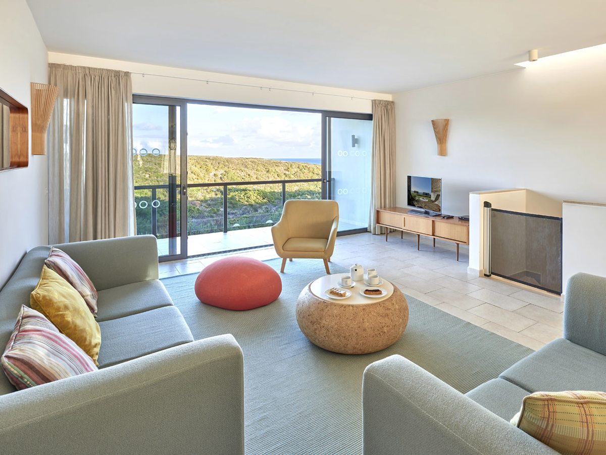 An upstairs living room with grey sofas, a round cork table and a balcony over scrubland to the sea, a mesh safety gate fitted at the top of the stairs.